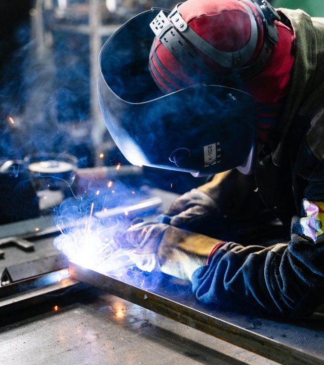 A welder works on metal fabrication indoors, creating bright sparks and smoke.